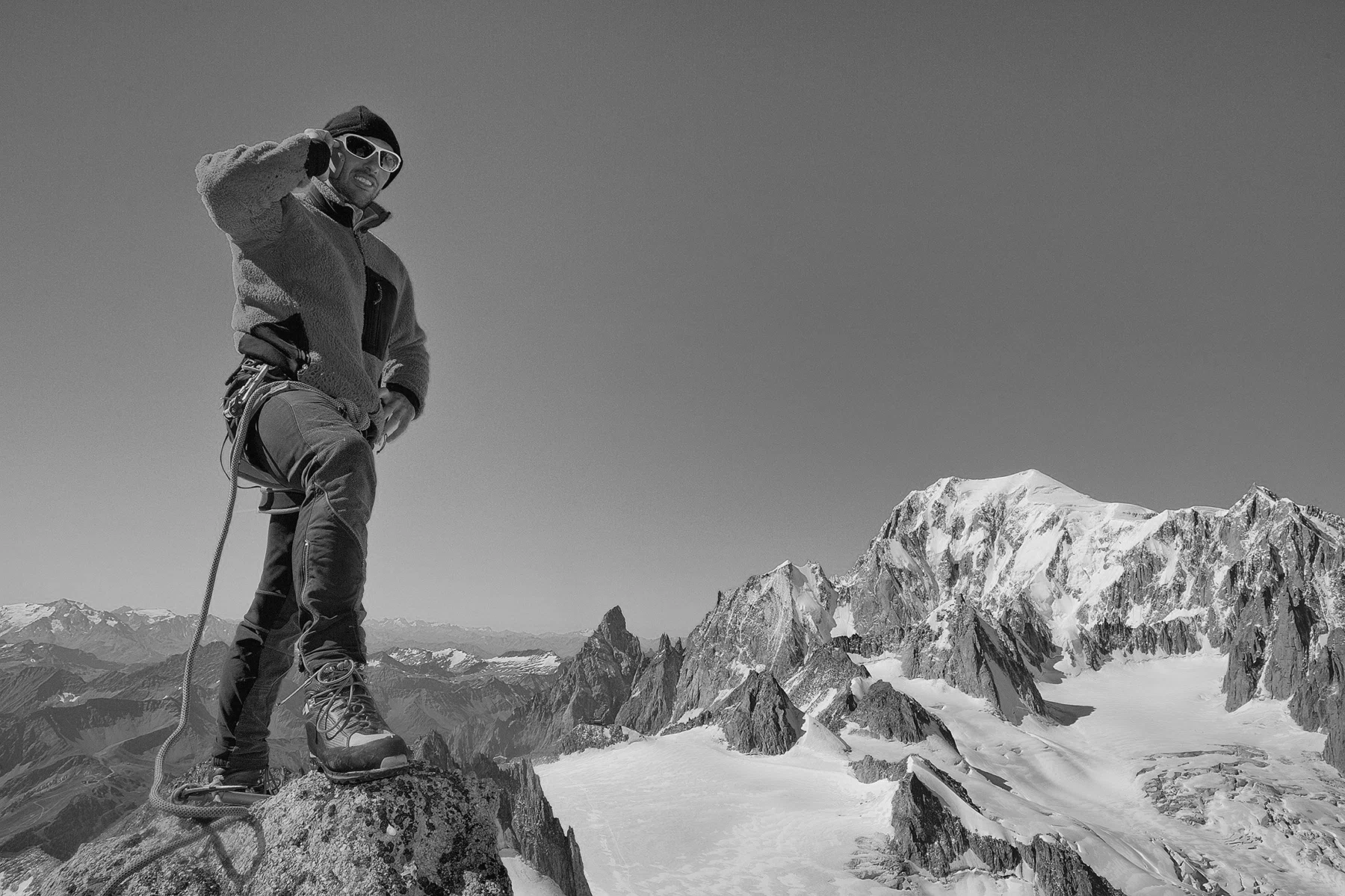 Climber standing on a rocky peak with mountain landscape and glaciers in the background, wearing winter climbing gear, sunglasses, and a fleece jacket.